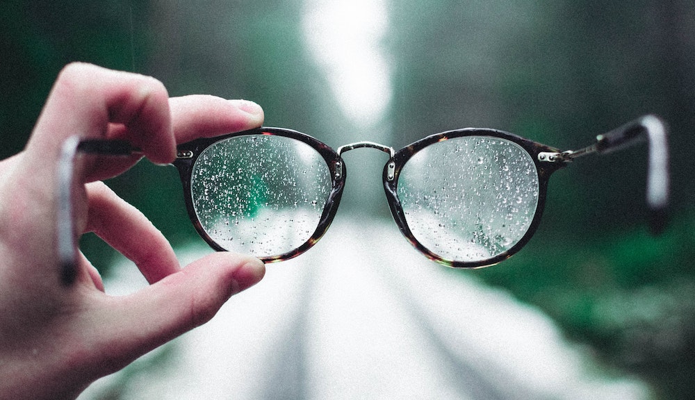 A hand holding eyeglasses spotted with rain and held out with a road disappearing into the woods behind them.